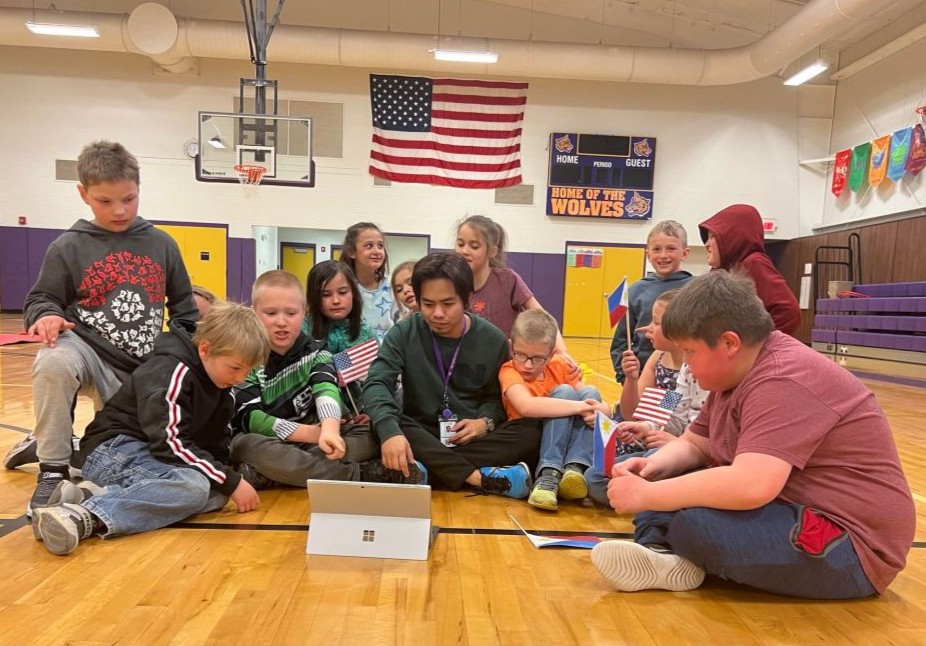 International teacher sat on floor surrounded by students viewing a laptop together